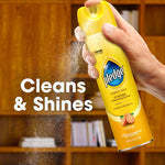 Hand spraying a yellow Pledge aerosol can, visible spray mist and text ‘Cleans & Shines’ over a blurred wood bookshelf background.