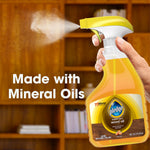 Hand spraying Pledge wood oil trigger bottle with visible mist and text stating made with mineral oils over wood shelving