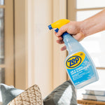 Hand holding a Zep Air & Fabric Odor Eliminator spray bottle aimed toward a couch, visible mist spraying from nozzle.