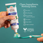 Hand holding a Guru Nanda toothpaste tube dispensing pale green paste onto a teal toothbrush with clean-ingredients icons.