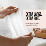 Hands stretching a single white wipe to show its extra-large, soft texture with product packaging blurred in the background.