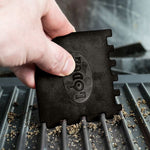 Hand holding a black Lodge multi-tooth scraper to clean debris from a ridged grill surface, logo visible.