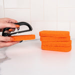 Hand holding a black scrub handle pressing an orange scouring pad, extra pads stacked nearby on counter.
