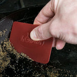 Hand using a red Lodge silicone scraper to remove debris from a cast-iron skillet, logo visible on scraper.