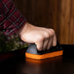 Person's hand gripping a black handle scrubber while pressing an orange scouring pad on a griddle surface.