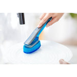 A hand washing a white plate with a blue Scotch-Brite dishwand under running water in a bright kitchen setting.