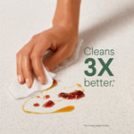 Hand wiping a sticky countertop with a textured white wipe and text claim stating "Cleans 3X better" against a neutral surface.