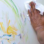 Hand using a cloth to wipe colorful paint and crayon marks from a white wall.
