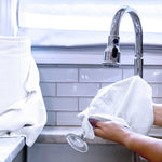 Hands drying a stemmed glass with a white microfiber towel in a kitchen sink scene