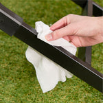 Hand holding a white pre-moistened cleaning cloth while wiping a black metal grill frame over grass.