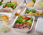Several Handi-foil aluminum meal prep pans filled with salads and entrees arranged on a light wood surface.