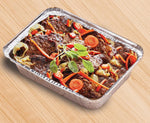 Aluminum meal prep pan filled with roasted meat and vegetables on a wood surface, shown at an angle