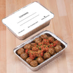 Close-up of a lidded Handi-foil aluminum pan labeled 'CONTAINS' beside an open pan filled with meatballs and sauce.