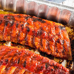 Barbecue-glazed ribs cooking in a disposable aluminum pan showing browned sauce and pan ridges