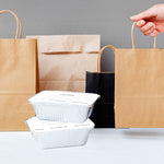 Two foil meal prep pans with folded lids stacked on a table in front of several paper shopping bags and a hand holding a bag handle