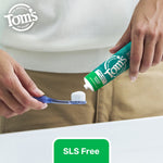 Close-up of hands squeezing Tom's of Maine Wicked Fresh toothpaste onto a toothbrush over a sink, with 'SLS Free' text below.
