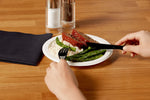 Hands using a black plastic knife and fork to cut meatloaf with asparagus and mashed potatoes on a paper plate
