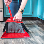 Hands holding the red broom handle while sweeping debris into the matching red dustpan, showing bristles and dustpan lip