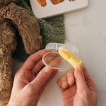 Hands holding a clear storage case and a yellow silicone finger toothbrush over a textured surface and toy