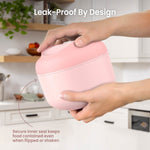 Hands holding a closed blush pink insulated bowl in a kitchen setting with text noting leak-proof design.