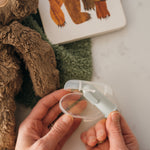 Hands holding a clear plastic case with a gray-green silicone finger toothbrush inside, soft toys and book blurred in the background.