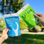 Hands holding two Grin pouches outdoors: a blue Double pouch and a green Clean pouch against blurred park background