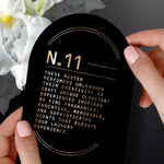 Hands holding a black card labeled ‘N.11’ with descriptive text about the perfumers, set against white flowers.