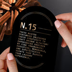 Hands holding black arch-shaped card labeled N.15 with descriptive copy, cinnamon sticks in the background