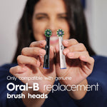 Hands holding two Oral‑B replacement brush heads (black and white variants) with overlay text stating they are genuine Oral‑B replacement brush heads.