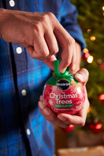 Hands holding and opening a Miracle-Gro for Christmas Trees bottle in front of a decorated tree.