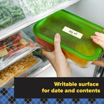 Hands placing a green-lidded container labeled 'July 1st' on a freezer shelf with bags of frozen vegetables visible.