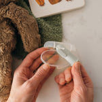 Hands placing a light green silicone finger toothbrush into a clear oval storage case near plush toys and a book