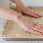 Close-up of hands smoothing unbleached parchment paper inside a rimmed baking sheet