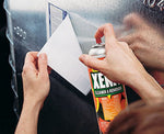 Hands holding a Stoner XENIT aerosol can and spraying to remove a paper sticker from a metal surface.