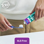Close-up of hands squeezing Tom's of Maine toothpaste onto a toothbrush above a sink with a purple "SLS Free" label at the bottom.