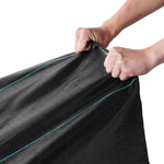 Close-up of two hands stretching a black woven landscape fabric showing texture and turquoise guide stitching