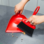 Hands using a red whisk broom and dustpan to sweep crumbs from a gray countertop