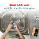 Top-down view of hands washing a dish in a stainless steel sink with an S.O.S box and pads visible at the side.