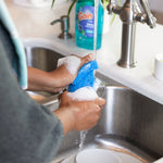 Hands scrubbing a glass at a kitchen sink with soapy sponge and a blurred Gain dish soap bottle on the counter