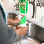 Hands scrubbing a glass with a green sponge and suds at a sink, a bottle of Gain dish soap visible in the background