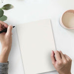 Overhead lifestyle view of hands holding and writing on a blank sheet of white paper beside a cup and green leaves