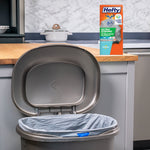 Kitchen scene showing a gray trash can lined with a Hefty Ultra Strong trash bag and a box of Hefty bags on the counter behind it