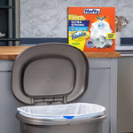 Kitchen scene with an open grey trash can lined with a white Hefty bag and Hefty product box on the counter behind it