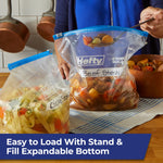 Kitchen scene showing two Hefty freezer bags standing upright filled with soup and stew, one being ladled into.