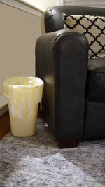 Yellow small trash can fitted with a yellow-and-white striped liner next to a dark leather sofa, showing bag draped over rim.