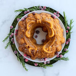 Golden holiday wreath cake on a white plate surrounded by rosemary sprigs and sugared cranberries