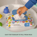 Hand pulling a wipe from an Honest Little Flushables pack with blue lid placed on a white surface with bath toys nearby