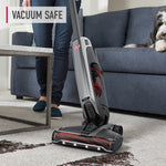 Upright Hoover vacuum cleaning a carpeted floor with a sofa and dog visible in the background
