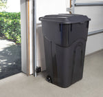 Black wheeled trash can with closed lid placed inside a garage near an open doorway, lifestyle setting.