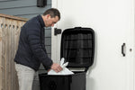 Lifestyle image of a person placing a tied trash bag into a black Hyper Tough wheeled garbage can with the lid open.